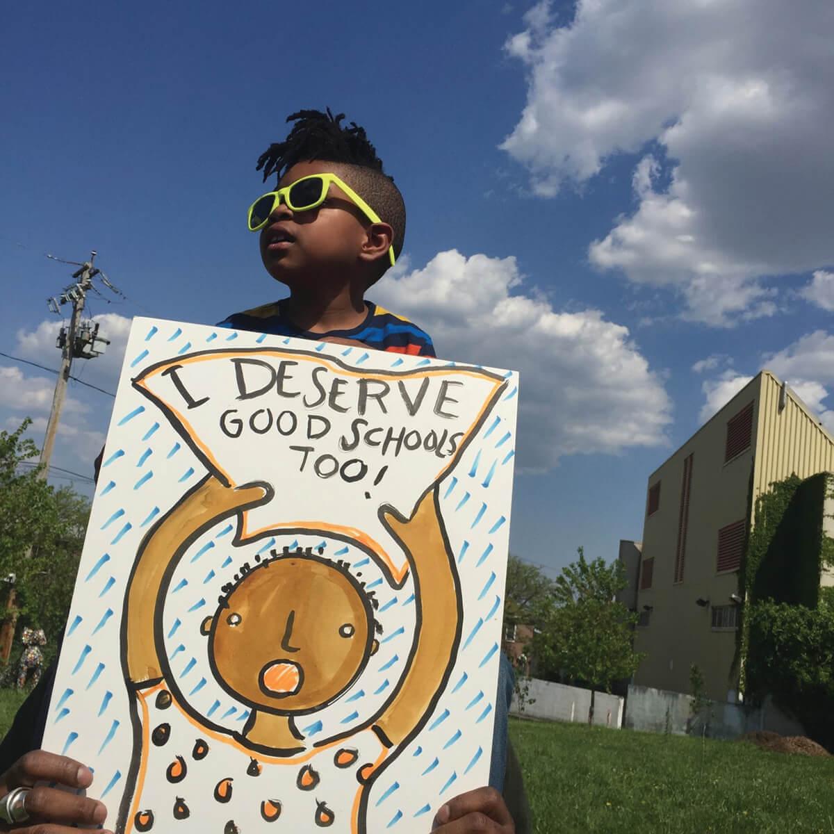 A young child holding a I deserve good schools too sign.