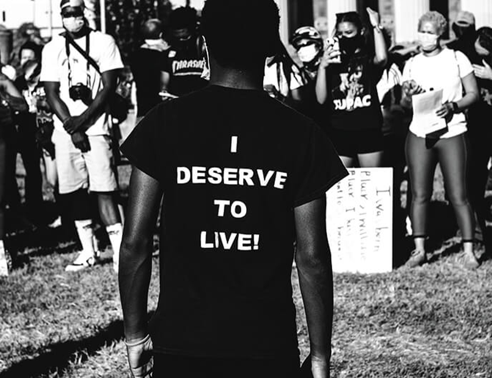 A young man facing towards a crowd in front of Baltimore City Hall with the words I deserve to live on the back of his shirt.